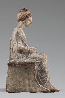 Figurine of a woman. A girl sitting in a chair is dressed in a soft white chiton over which a blue,