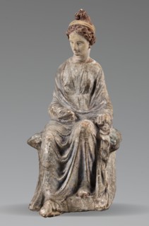 Figurine of a woman. A girl sitting in a chair is dressed in a soft white chiton over which a blue,