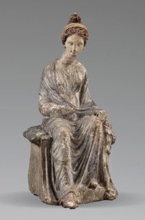 Figurine of a woman. A girl sitting in a chair is dressed in a soft white chiton over which a blue,