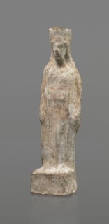 Female terracotta figurine of a standing woman, depicted frontally. Set on a small, hollow,