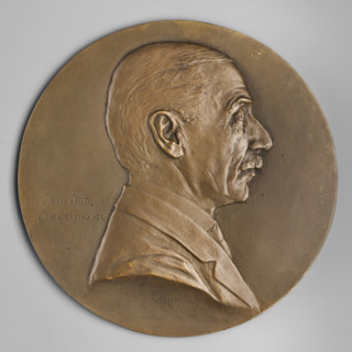 Bust of a man in right profile, hair smoothly combed back, mustache trimmed, jacket on the