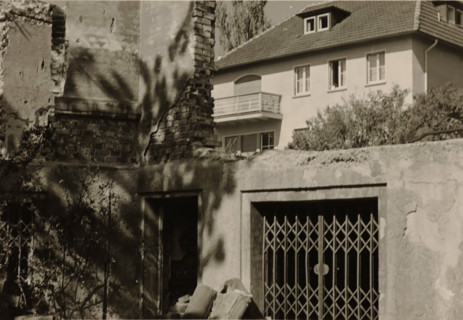 Part of a ruin with a sliding gate and a doorway in it, with a large house with a balcony in the