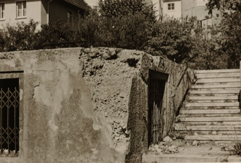 Part of a ruin with a sliding gate and a doorway in it, with a large house with a balcony in the