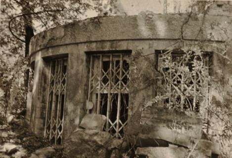 Circular part of a ruin, with bars in front of the windows, stones in the foreground and vegetation