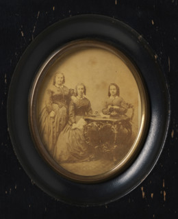 Group portrait of three unknown women around a table, with photos on top, frame, photograph, label: