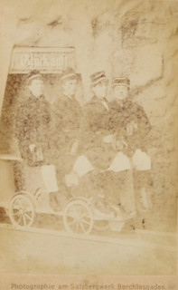 Group portrait of four women wearing caps, sitting on a carriage on a rail, cabinet photograph,