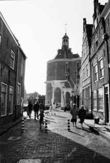 Rue pavée bordée de maisons de briques, avec une tour au fond et des piétons en contre-jour.