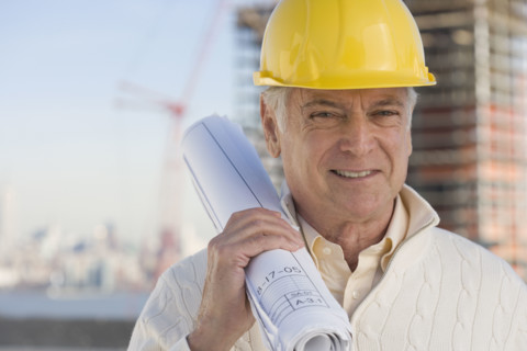 Senior man wearing hard hat and holding blueprints.