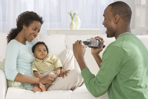 Father video recording mother and baby on sofa.