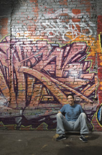 Asian man sitting in front of graffitied wall. Date : 2007
