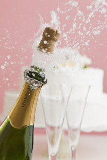 Cork exploding from champagne bottle with wedding cake in background.