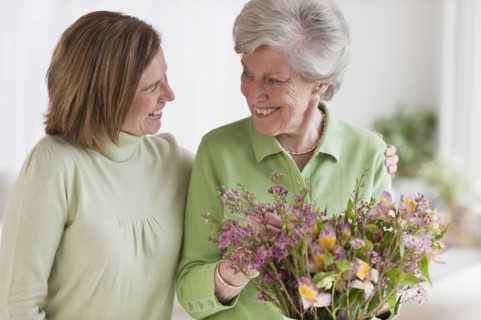 Daughter hugging mother holding flowers.
