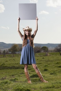 Cowgirl holding up blank poster while standing in field. Photo : Mike Kemp