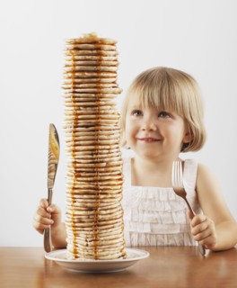 Young girl sitting behind a tall stack of pancakes. Photo. FBP