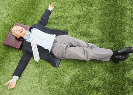Happy businessman lying on lawn with briefcase under head. Photo : Jamie Grill