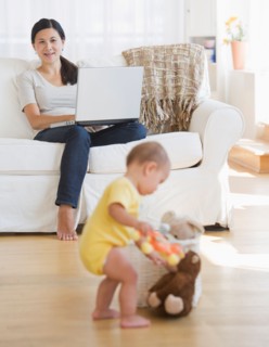 Baby boy (6-11 months) playing, mother sitting on sofa and using laptop. Photo : Daniel Grill