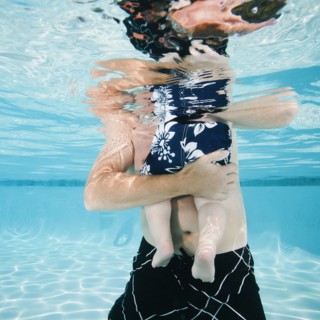 Father and son (12-18months) standing in swimming pool.