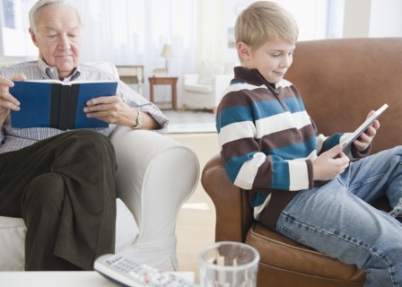 Grandfather reading book, grandson (8-9) reading e-book in living room. Photo : Jamie Grill