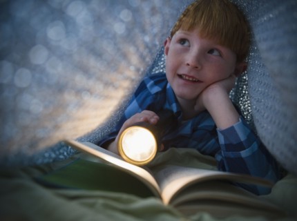 Boy (8-9) reading book under bed covers. Photo : Jamie Grill Photography