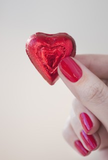 Close up of woman's fingers with red nail polish holding chocolate in shape of heart in red wrapping.