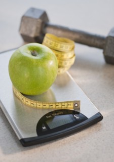 Green apple on weight scale, tape measure and exercise weight in background.