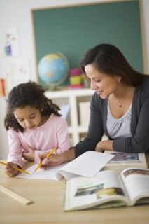 Girl (6-7) with female teacher sitting at desk in classroom.