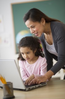 Girl (6-7) with female teacher using laptop in classroom.