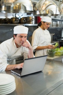 Chef using laptop and cook preparing food in commercial kitchen.