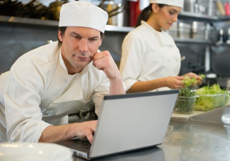 Chef using laptop and cook preparing food in commercial kitchen.