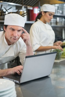 Chef using laptop and cook preparing food in commercial kitchen.