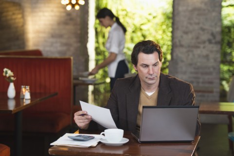 Owner working with laptop in restaurant with waitress in background.