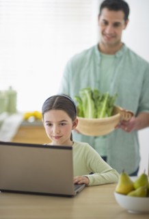 Girl (8-9) using laptop and father preparing food in domestic kitchen.