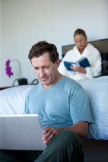 Mature man with laptop in bedroom and woman reading book in bed. Photo: Rob Lewine