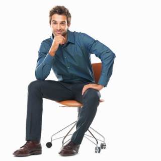 Studio shot of young business man sitting on chair with hand on chin and smiling. Photo: