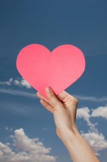Woman's hand holding pink heart shape against sky. Photo: Winslow Productions