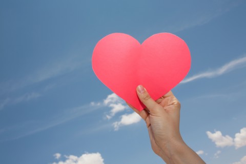 Woman's hand holding pink heart shape against sky. Photo: Winslow Productions