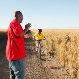 Father clapping as son (8-9) is cycling along dirt road in fields.