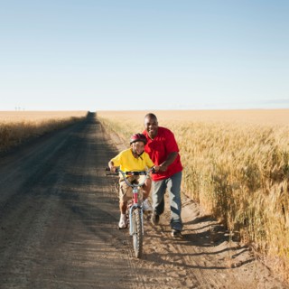 Father teaching son (8-9) how to cycle on along dirt road.