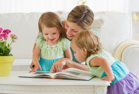 Mother with daughters (2-3) reading book in living room.