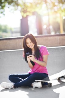 Young chinese woman text messaging while sitting on skateboard. Photo : Take A Pix Media