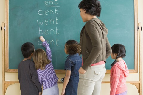 Children (6-7) writing on blackboard with teacher looking at them. Photo: Rob Lewine