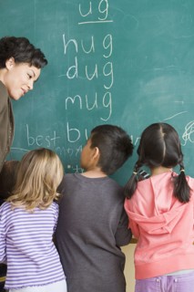 Children (6-7) writing on blackboard with teacher looking at them. Photo: Rob Lewine
