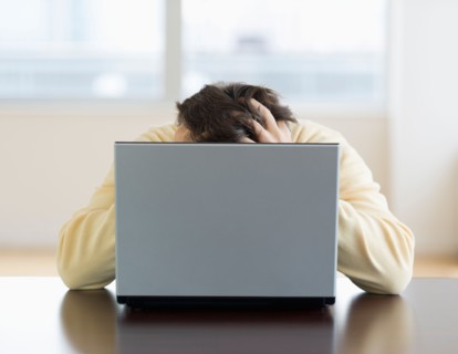 Stressed man sitting at desk behind laptop with head in hands.