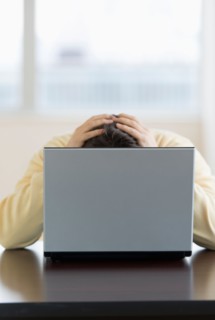 Stressed man sitting at desk behind laptop with head in hands.