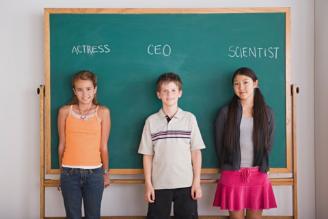 Two schoolgirls and schoolboy standing against blackboard with profession names written above.