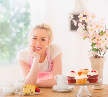 Smiling young woman in apron leaning on table with cakes. Photo : Daniel Grill