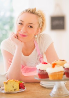 Smiling young woman in apron leaning on table with cakes. Photo : Daniel Grill