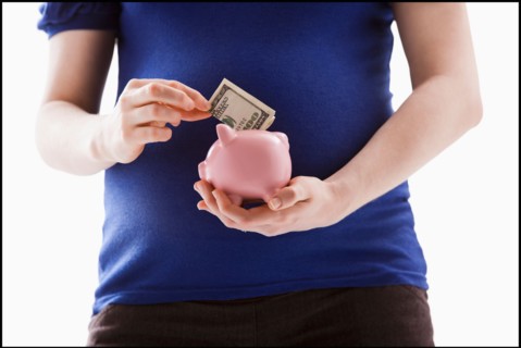 Studio Shot of mid section of pregnant woman putting money into Piggy Bank. Photo : Mike Kemp