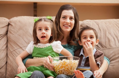 Front view of mother with children (4-5, 6-7) sitting on couch watching tv and eating popcorn.