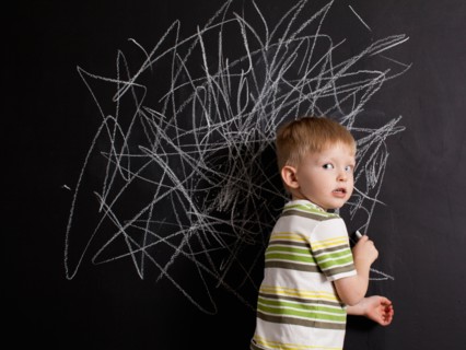Cute toddler boy (2-3) standing against blackboard with daub written in chalk. Photo : Jessica